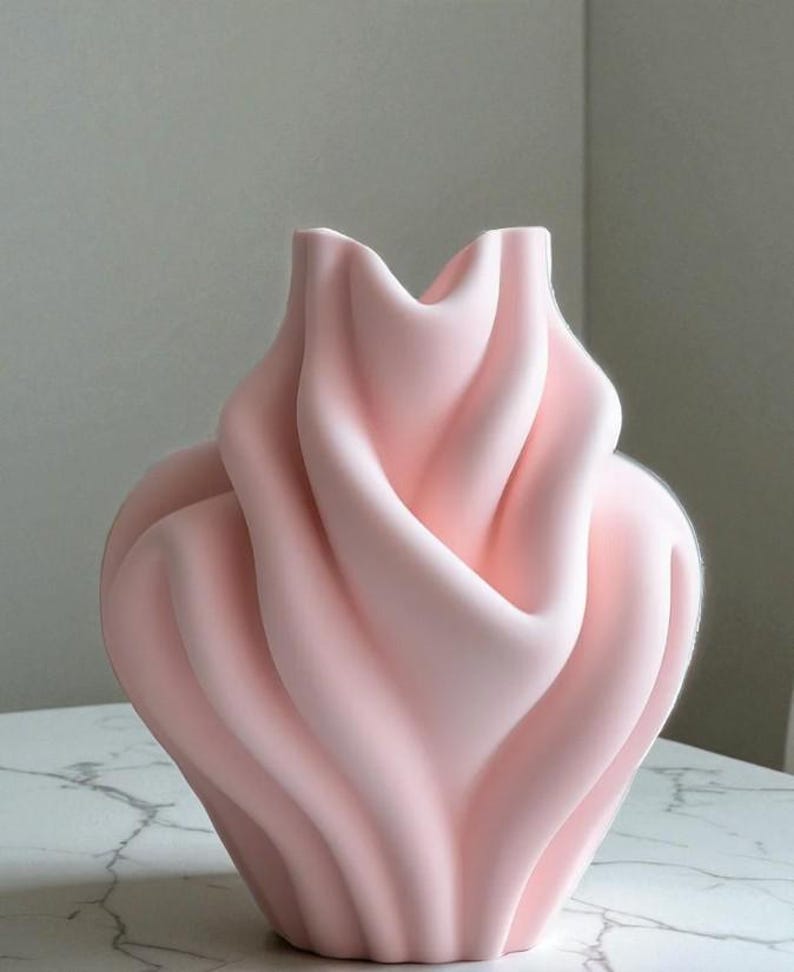 May include: A light pink ceramic vase with a unique, flowing design. The vase has a wide, rounded base that tapers upwards, with the top opening in a wavy, petal-like shape. The smooth, matte finish adds to its modern aesthetic.