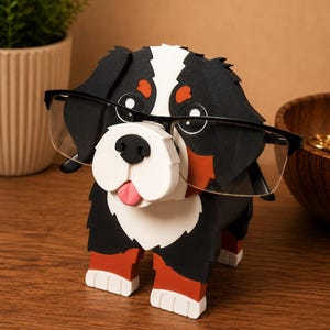 May include: A Bernese Mountain Dog-shaped glasses holder. The holder is black, white, and brown with a pink tongue and red accents on the feet. The dog is wearing black-framed glasses. A decorative item for eyeglasses.