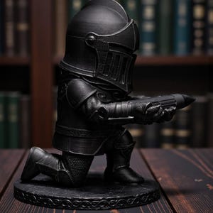 May include: A black figurine of a knight kneeling on a round base, holding a long, cylindrical object. The knight is wearing full armor, including a helmet with a visor. The base has a decorative pattern. The background is blurred, with bookshelves.