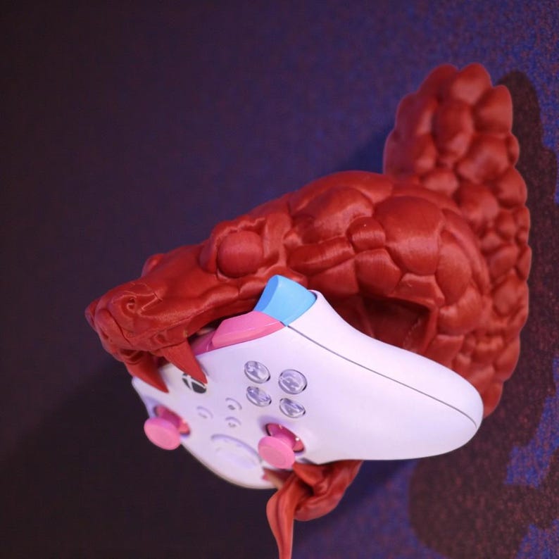 May include: A red, 3D-printed snake head wall mount holding a white and pink video game controller. The controller has pink and blue accents on the buttons and thumbsticks. The snake's mouth is open, and the controller is held in its jaws.