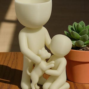 May include: A cream-colored planter shaped like a person holding a dog, with another person kneeling nearby. A small succulent plant in an orange pot is in the background. The planter has a large head and a small body.