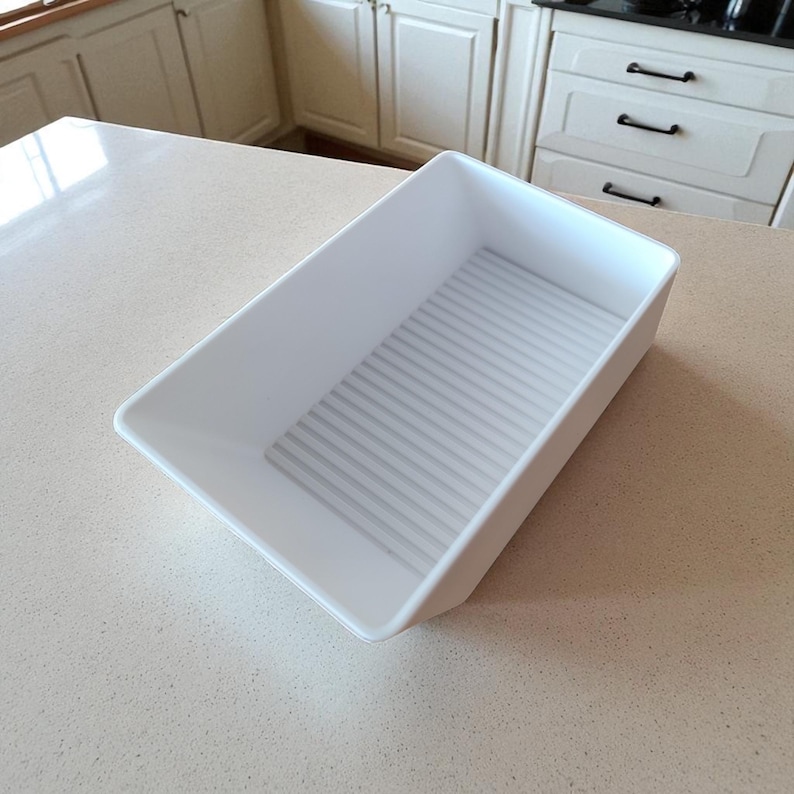 May include: A white rectangular plastic storage bin with a ridged bottom. The bin is empty and angled, showing its interior. The bin is on a light-colored countertop, likely in a kitchen setting.