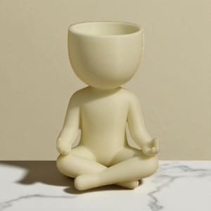 May include: A cream-colored figurine of a person in a seated yoga pose. The head is a large, open bowl shape. The figure is sitting cross-legged with hands resting on the knees. The figurine is on a white marble surface.