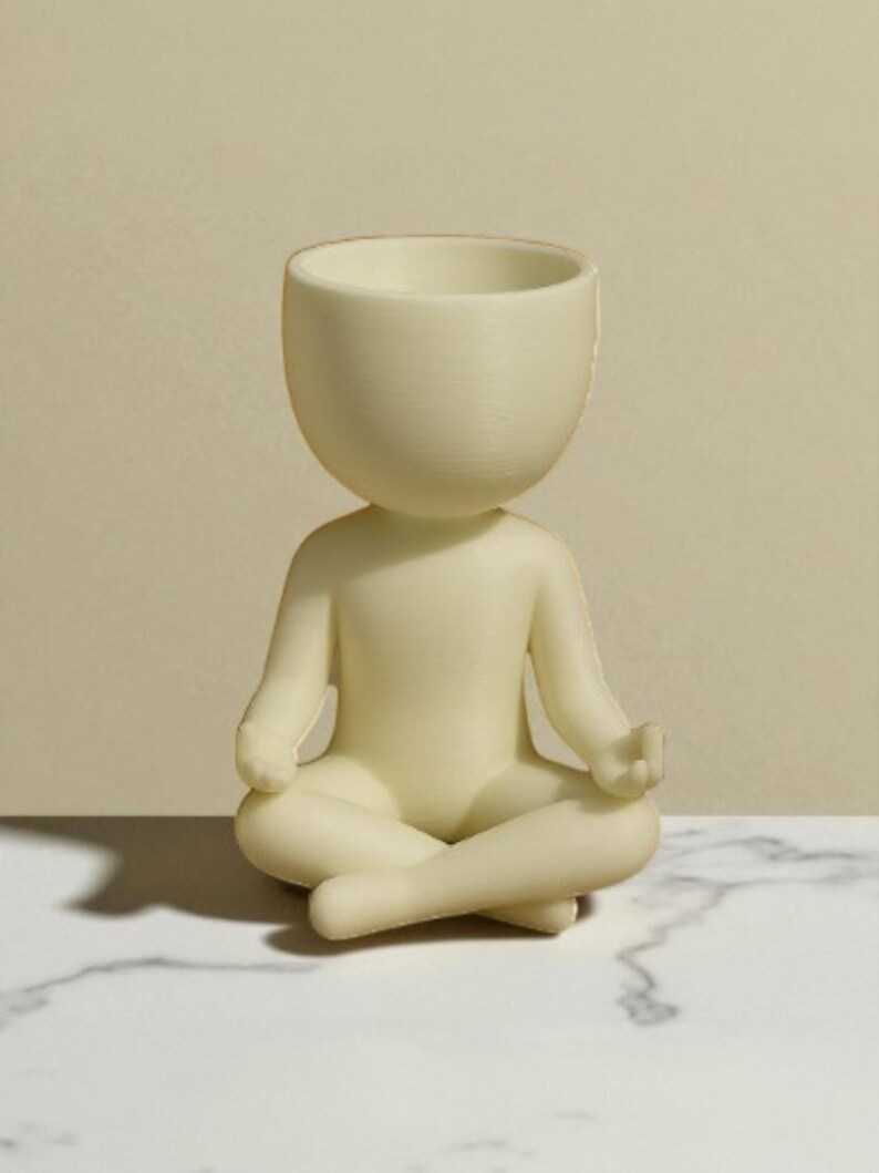 May include: A cream-colored figurine of a person in a seated yoga pose. The head is a large, open bowl shape. The figure is sitting cross-legged with hands resting on the knees. The figurine is on a white marble surface.