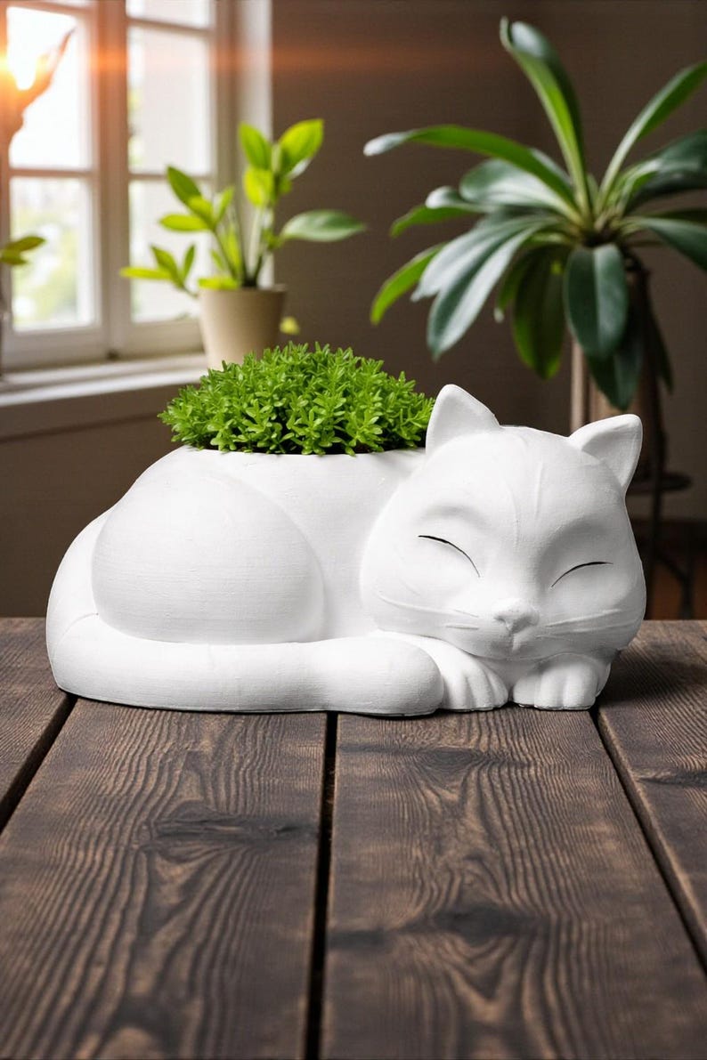 May include: A white ceramic cat-shaped planter, with a sleeping cat design. The planter is filled with green plants. The cat planter is on a dark wooden surface, with a window and plants in the background.