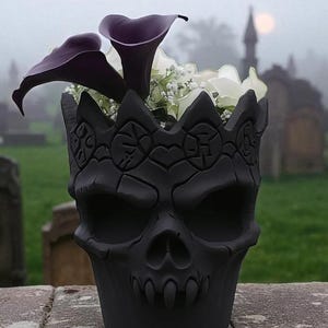 May include: A black skull-shaped planter with a crown design, filled with purple calla lilies and white flowers. The planter is set against a blurred background of a cemetery on a foggy day.