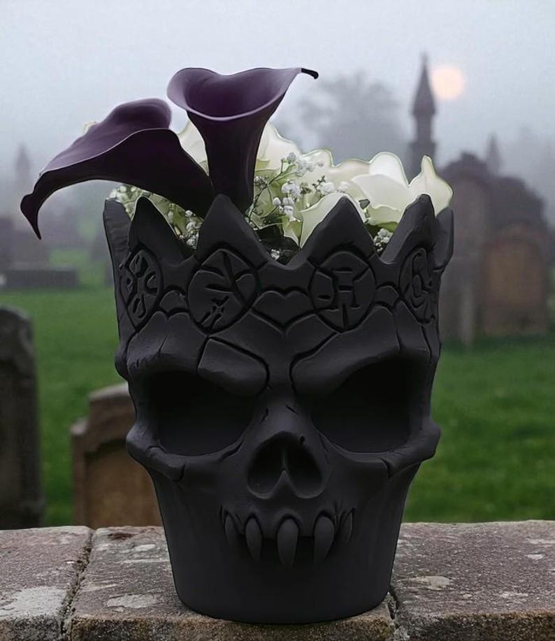 May include: A black skull-shaped planter with a crown design, filled with purple calla lilies and white flowers. The planter is set against a blurred background of a cemetery on a foggy day.
