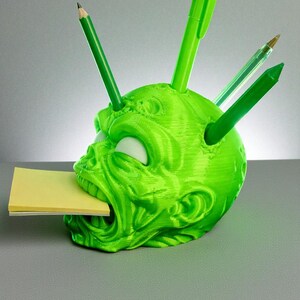 May include: A neon green, 3D-printed desk organizer shaped like a zombie head. The head has white eyes, a wide open mouth holding a yellow sticky note pad, and holes in the head for pens and pencils. Several green pens and pencils are inserted into the head.