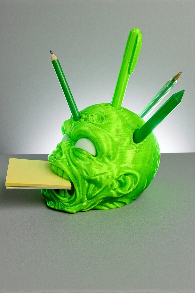 May include: A neon green, 3D-printed desk organizer shaped like a zombie head. The head has white eyes, a wide open mouth holding a yellow sticky note pad, and holes in the head for pens and pencils. Several green pens and pencils are inserted into the head.
