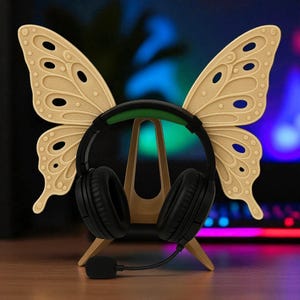 May include: A black headset with a microphone sits on a wooden butterfly-shaped stand. The beige butterfly wings have detailed cutouts. The headset features a green band and black earcups.