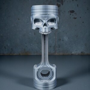 May include: A silver-colored piston with a skull-shaped head. The skull has detailed eye sockets, a nose cavity, and a row of teeth. The piston's connecting rod and base are also visible, creating a unique decorative piece.