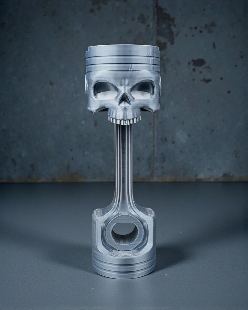 May include: A silver-colored piston with a skull-shaped head. The skull has detailed eye sockets, a nose cavity, and a row of teeth. The piston's connecting rod and base are also visible, creating a unique decorative piece.