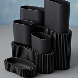 May include: A collection of black, oval-shaped ceramic vases and containers. The vases are arranged in a tiered display, with some resting on ribbed platforms. The matte finish and minimalist design create a modern aesthetic.