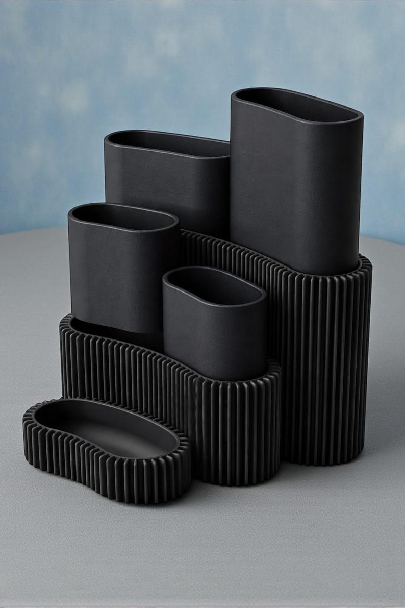 May include: A collection of black, oval-shaped ceramic vases and containers. The vases are arranged in a tiered display, with some resting on ribbed platforms. The matte finish and minimalist design create a modern aesthetic.