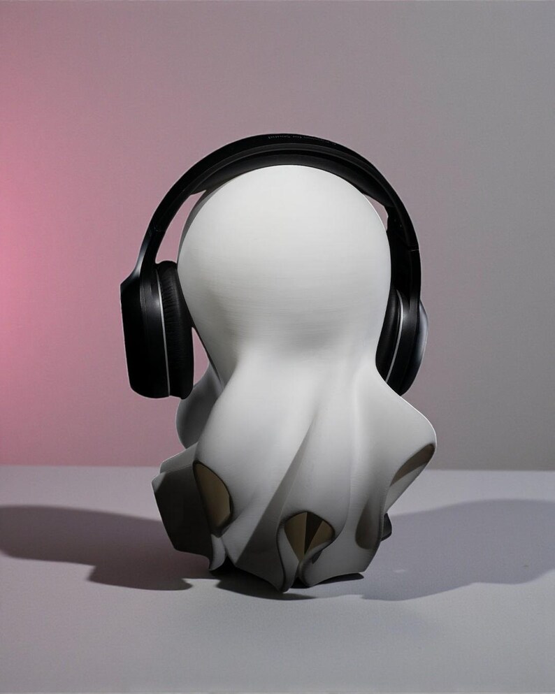May include: A white sculpture of a ghost-like figure wearing black headphones. The figure has a rounded head and a flowing, draped body with dark accents. The background transitions from pink to gray.