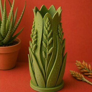 May include: A green, decorative vase with an aloe vera plant design. The vase has a textured surface and is shaped like a tall, slender cylinder. The design includes detailed depictions of aloe vera leaves and flower stalks. The vase is approximately 8 inches tall.