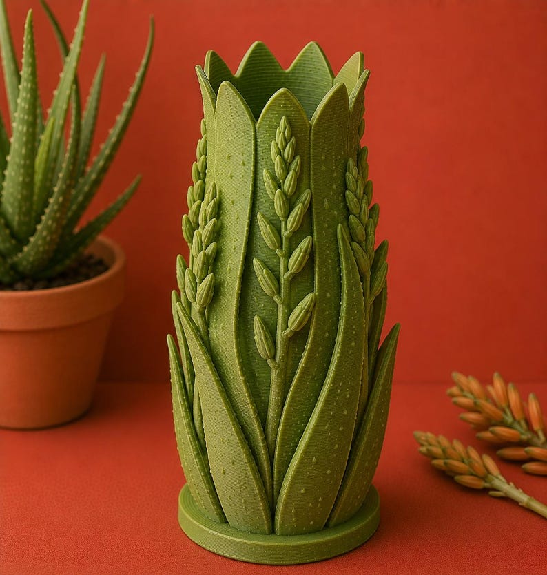 May include: A green, decorative vase with an aloe vera plant design. The vase has a textured surface and is shaped like a tall, slender cylinder. The design includes detailed depictions of aloe vera leaves and flower stalks. The vase is approximately 8 inches tall.