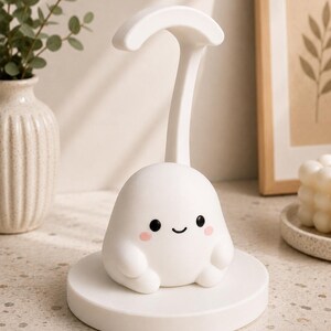 May include: A white, cartoon-like night light with a smiling face, rosy cheeks, and small arms. The light sits on a round base and has a curved handle. The lamp is designed to look like a friendly ghost.