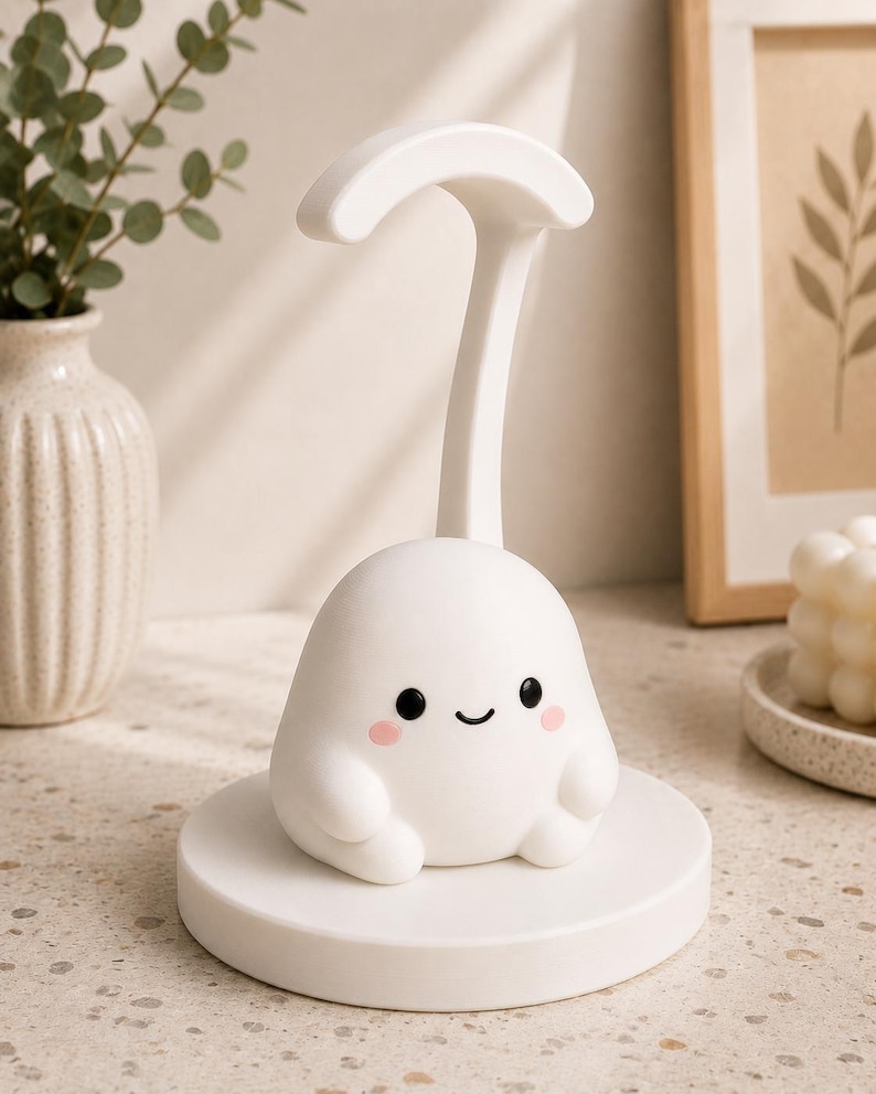 May include: A white, cartoon-like night light with a smiling face, rosy cheeks, and small arms. The light sits on a round base and has a curved handle. The lamp is designed to look like a friendly ghost.