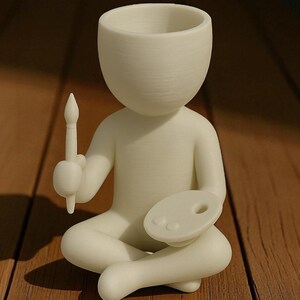 May include: A cream-colored figurine of a person sitting cross-legged, holding a paintbrush and a palette. The figurine has a large head with a hollowed-out top, possibly for a plant. The figure is on a wooden surface.