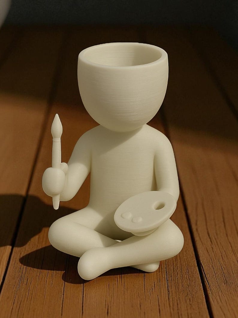 May include: A cream-colored figurine of a person sitting cross-legged, holding a paintbrush and a palette. The figurine has a large head with a hollowed-out top, possibly for a plant. The figure is on a wooden surface.