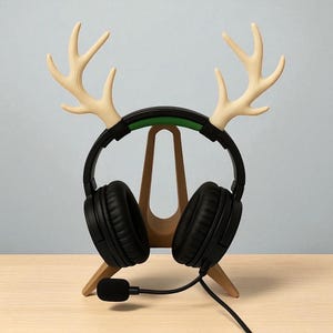 Op de afbeelding: Een zwarte headset met een microfoon rust op een houten standaard in de vorm van een hertenkop. De standaard heeft lichtgekleurde geweien en een groene streep op de headset. De standaard staat op een lichtgekleurd houten oppervlak.