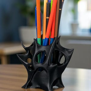 May include: A black, abstract-shaped pencil holder with a unique, spiky design. The holder contains a variety of colored pencils and pens, including orange, red, and blue. The holder sits on a wooden surface.