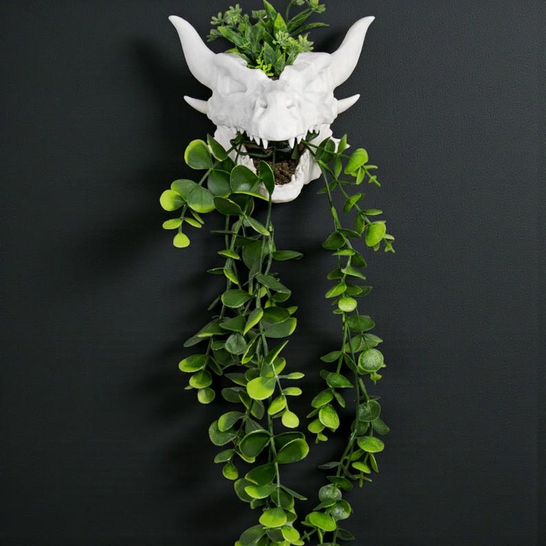Puede incluir: Una maceta de pared blanca con forma de cr&aacute;neo de drag&oacute;n, llena de plantas verdes. La maceta tiene cuernos y un dise&ntilde;o detallado de calavera. Las vides verdes colgantes caen en cascada, creando una pieza decorativa &uacute;nica. El fondo es negro liso.