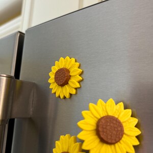 May include: Yellow sunflower magnets with brown centers are attached to a stainless steel refrigerator door. The magnets have a three-dimensional design, with detailed petals and a textured center. The magnets are decorative and add a touch of nature to the kitchen.