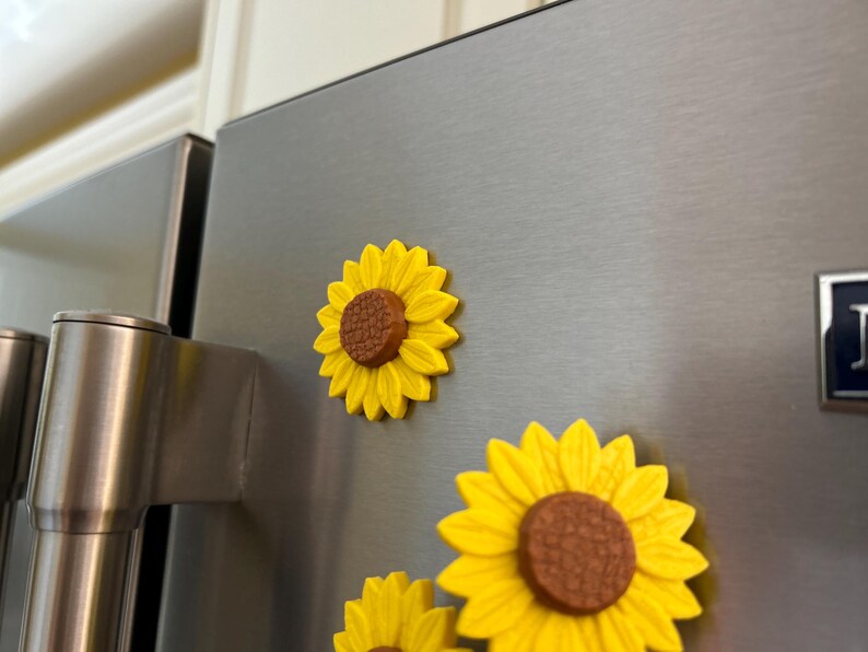 May include: Yellow sunflower magnets with brown centers are attached to a stainless steel refrigerator door. The magnets have a three-dimensional design, with detailed petals and a textured center. The magnets are decorative and add a touch of nature to the kitchen.