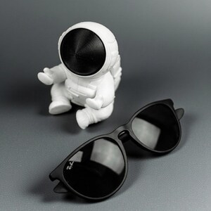 May include: A pair of black sunglasses with dark lenses and a white astronaut figurine on a gray surface. The sunglasses have a classic design with a subtle logo on the left lens.