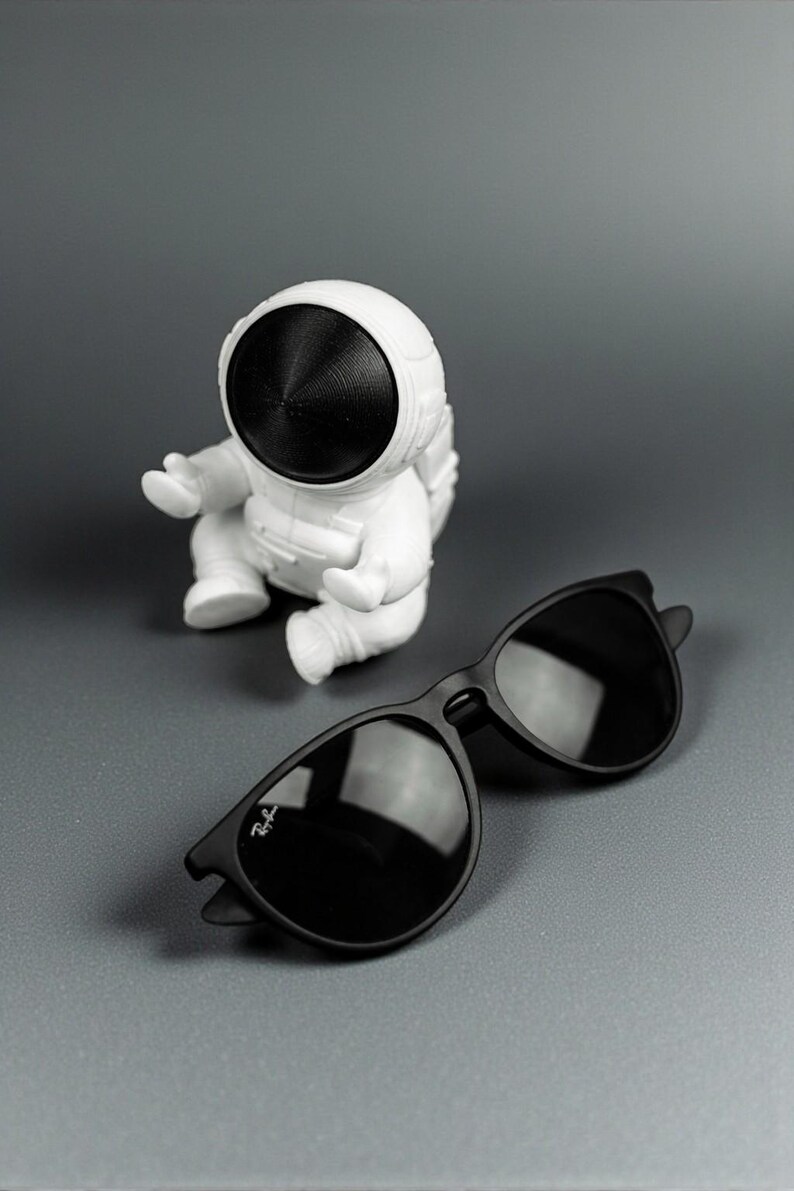 May include: A pair of black sunglasses with dark lenses and a white astronaut figurine on a gray surface. The sunglasses have a classic design with a subtle logo on the left lens.