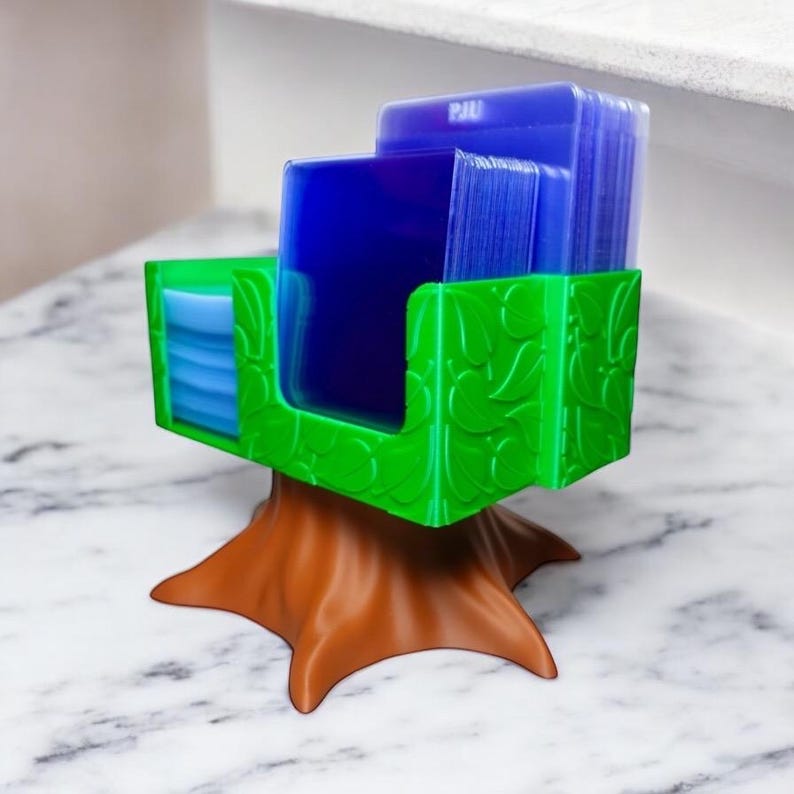 May include: A 3D-printed card holder in the shape of a tree. The holder has a brown trunk and a green top with leaf-like designs. Blue cards are visible inside the holder. The letters "PJU" are visible on the cards.
