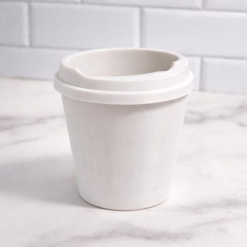May include: A white, reusable coffee cup with a lid. The cup has a textured surface and a curved handle on the lid. The cup is made of a durable material and is designed for hot or cold beverages.