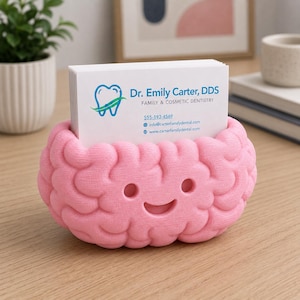 May include: A pink brain-shaped business card holder with a smiling face. The holder contains a stack of white business cards with the text "Dr. Emily Carter, DDS" and contact information. The holder is on a wooden surface.