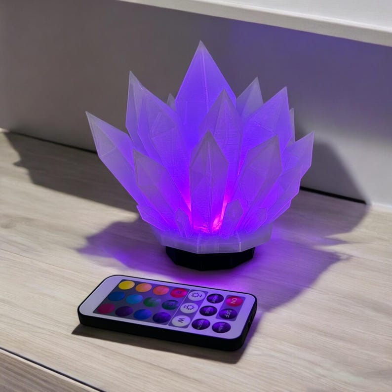 May include: A crystal-shaped lamp with a white exterior and a vibrant purple glow emanating from within. A remote control with color options sits in front of the lamp. The lamp is on a wooden surface.