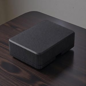 May include: A rectangular, matte black box with a textured surface and vertical ribbed detailing on the sides. The box is placed on a dark wooden surface, creating a contrast in tones. The design is minimalist and modern.