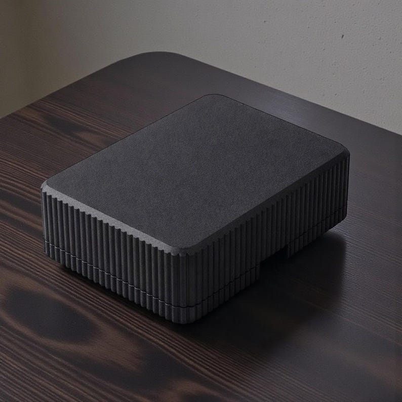 May include: A rectangular, matte black box with a textured surface and vertical ribbed detailing on the sides. The box is placed on a dark wooden surface, creating a contrast in tones. The design is minimalist and modern.