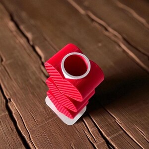 K&ouml;nnte beinhalten: Eine rote, abstrakte, 3D-gedruckte Skulptur mit einem wei&szlig;en Innenzylinder. Die Skulptur besteht aus gestapelten, gebogenen, bandartigen Formen mit diagonalen Linien. Sie steht auf einer kleinen, wei&szlig;en, rechteckigen Basis. Der Hintergrund ist eine Holzoberfl&auml;che.
