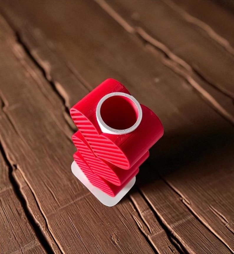 K&ouml;nnte beinhalten: Eine rote, abstrakte, 3D-gedruckte Skulptur mit einem wei&szlig;en Innenzylinder. Die Skulptur besteht aus gestapelten, gebogenen, bandartigen Formen mit diagonalen Linien. Sie steht auf einer kleinen, wei&szlig;en, rechteckigen Basis. Der Hintergrund ist eine Holzoberfl&auml;che.