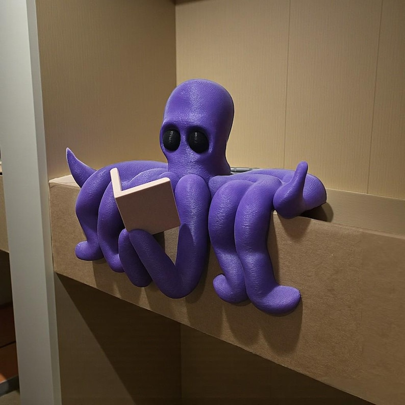 May include: A purple octopus figurine reading a small book. The octopus has large, black eyes and is perched on a shelf. The book is a light tan color. The figurine is a whimsical decorative item.