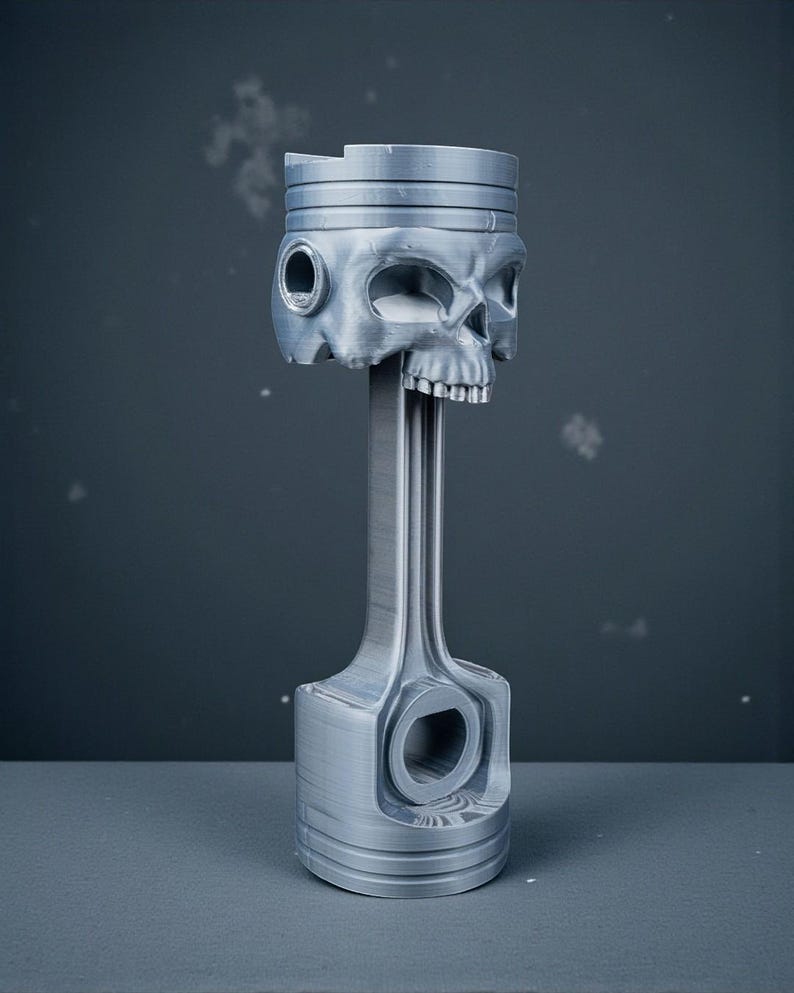 May include: A silver-colored piston with a skull-shaped head. The skull has detailed features, including eye sockets, a nose cavity, and teeth. The piston rod extends downward, connecting to a base with a circular opening.