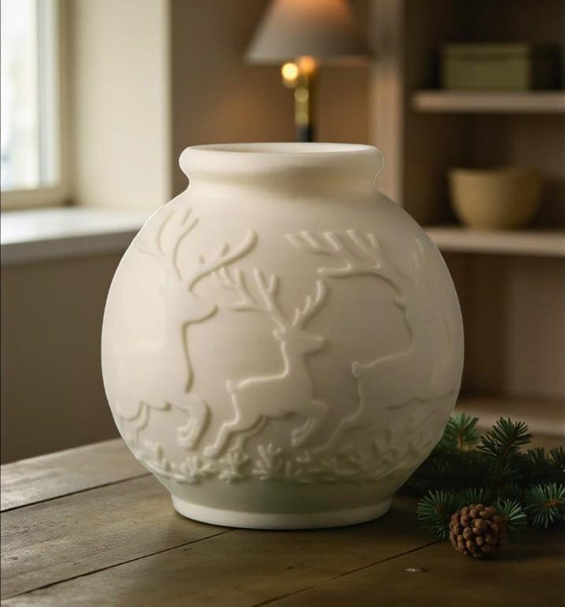 May include: Cream-colored ceramic vase with a round shape and a slightly flared rim. The vase features a raised relief design of deer in a forest setting. A pine cone and evergreen sprigs are on the wooden surface.