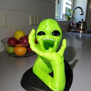 May include: A bright green alien figurine with large black eyes and an open mouth, mounted on a black base. The alien has one arm raised. A bowl of fruit is in the background.