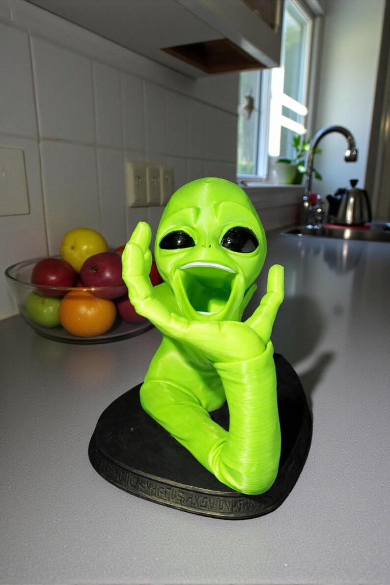 May include: A bright green alien figurine with large black eyes and an open mouth, mounted on a black base. The alien has one arm raised. A bowl of fruit is in the background.