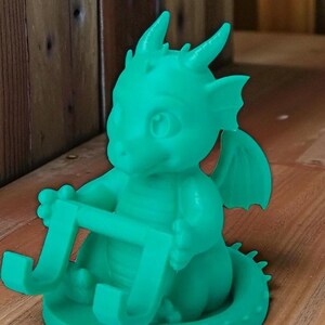 May include: A teal-colored dragon-shaped phone holder. The dragon has horns, wings, and is holding a rectangular frame. The holder is sitting on a wooden surface. The dragon is a whimsical, fantasy-themed item.