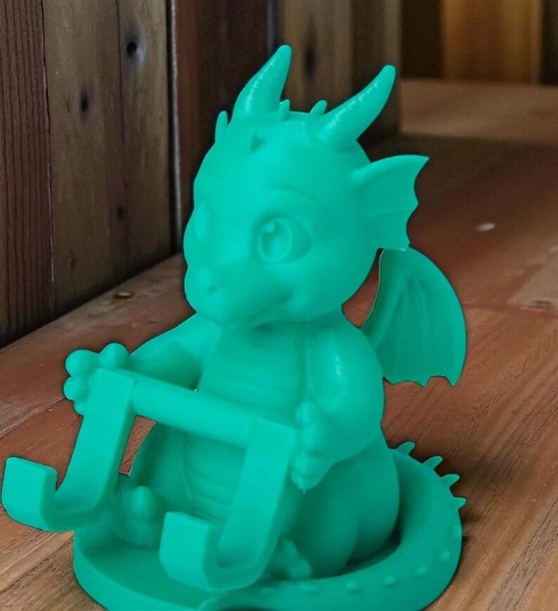 May include: A teal-colored dragon-shaped phone holder. The dragon has horns, wings, and is holding a rectangular frame. The holder is sitting on a wooden surface. The dragon is a whimsical, fantasy-themed item.