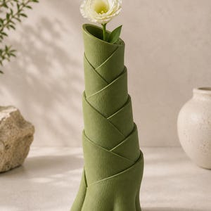 May include: A green, sculptural vase with a unique, layered design, holding a single white flower. The vase has a textured surface and a flared base. The flower has a delicate, multi-petaled bloom.
