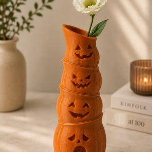 May include: An orange, stacked pumpkin vase with carved jack-o'-lantern faces. A white flower with a green stem and leaf is placed in the top opening. The vase is a decorative item for Halloween.
