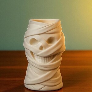 May include: A white ceramic tiki mug shaped like a mummy's head. The mug features a skull face with wrapped bandages, creating a spooky and unique design. The mug is designed for beverages and is suitable for Halloween or themed parties.